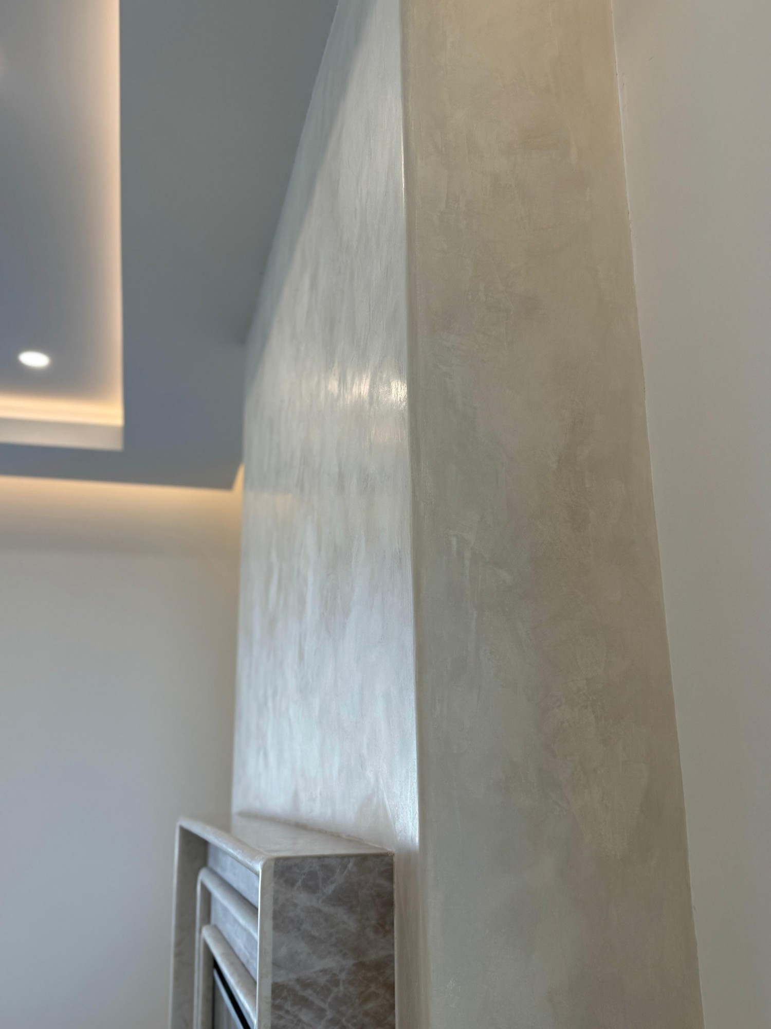 A chimney breast in pale Grassello Fino seen from the side, its polished face catching the light from a ceiling cove above a marble fireplace surround.