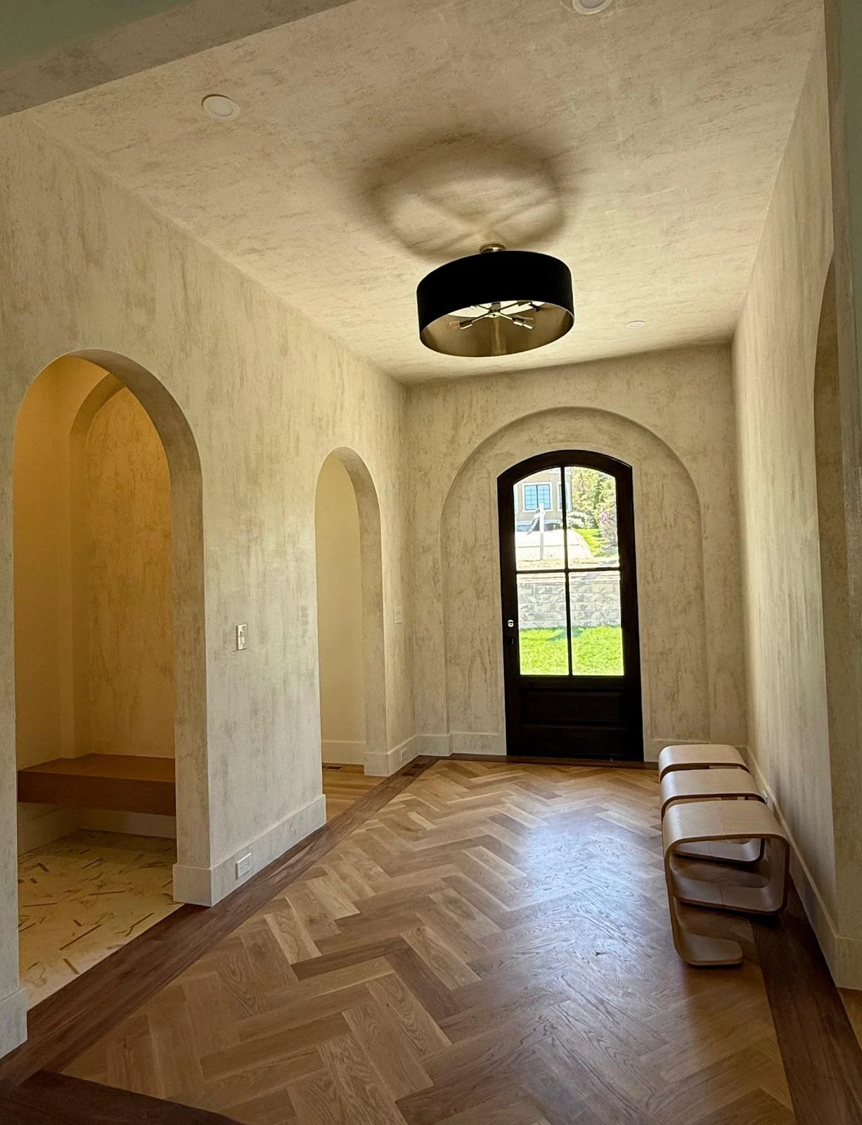 An entry hall finished in a travertine-effect plaster across the walls, the arched openings and the ceiling, with a herringbone oak floor and an arched front door.